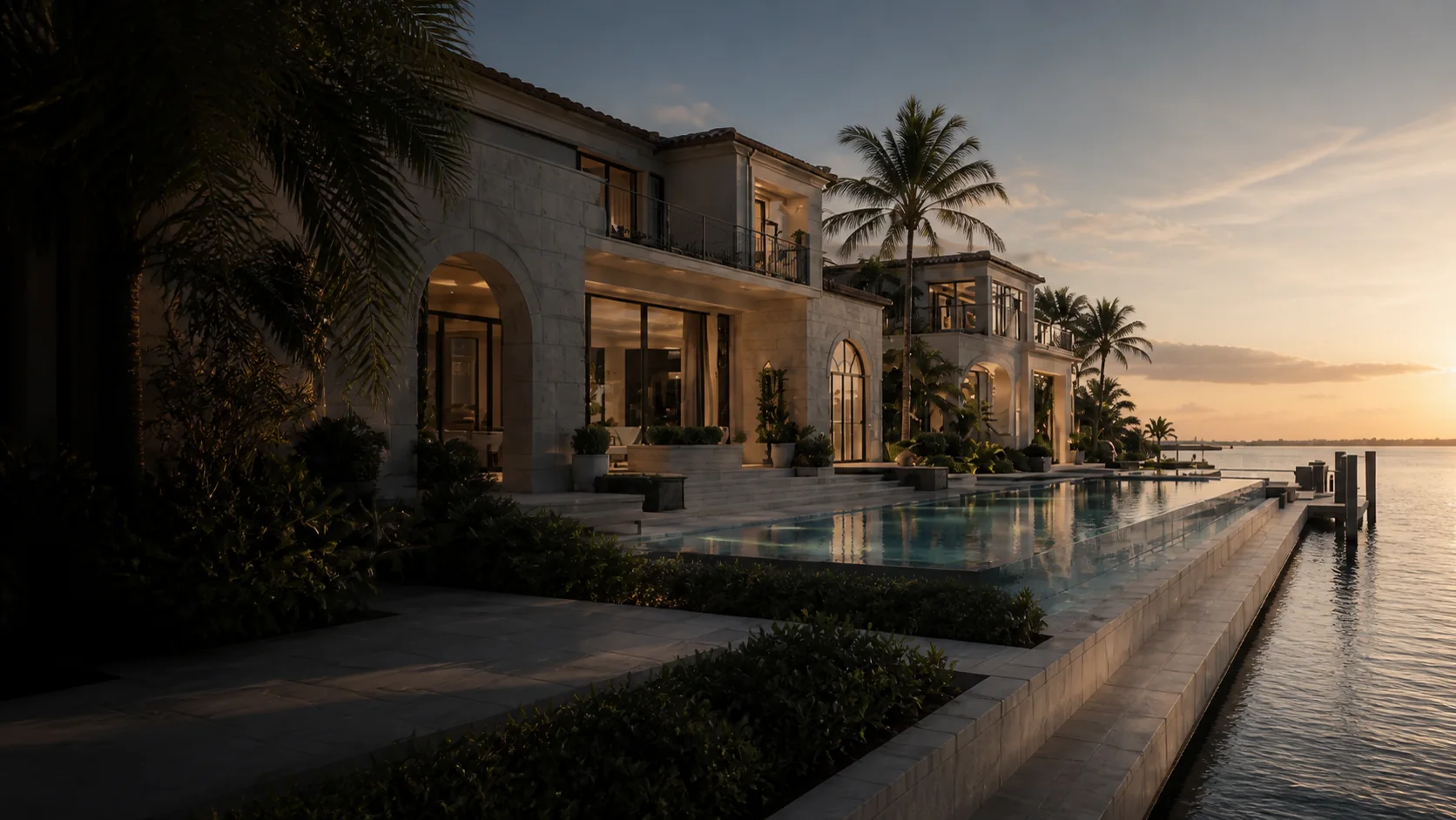 Waterfront residence with a limestone facade, illuminated rooms, palms, an infinity pool, and calm water at sunset.